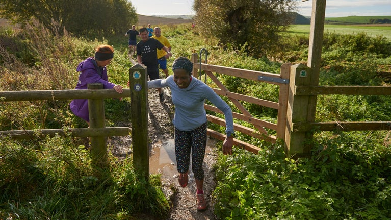 Runners on a track flanked by bushes running towards the camera the front one coming through a gate